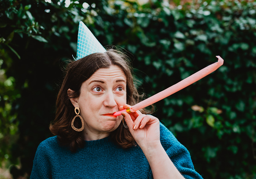 Leanne blows a party horn while wearing a party hat, standing in front of a bush. Leanne blows a party horn while wearing a party hat, standing in front of a bush.